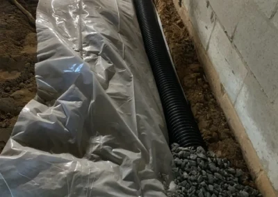 A construction area showing a black drainage pipe next to a clear plastic sheet and a layer of gravel, surrounded by dirt and concrete walls