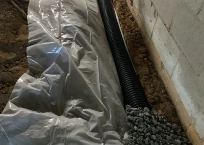 A construction site featuring a long plastic-covered trench with gravel and a black drainage pipe running alongside a concrete block wall