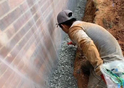 A construction worker in a gray shirt and cap is crouching in a narrow trench lined with gravel, surrounded by plastic sheeting
