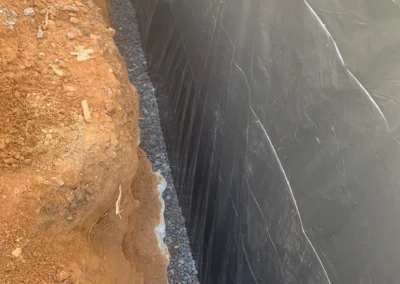 A deep trench lined with black plastic, filled with gravel, surrounded by reddish-brown earth, illuminated by sunlight