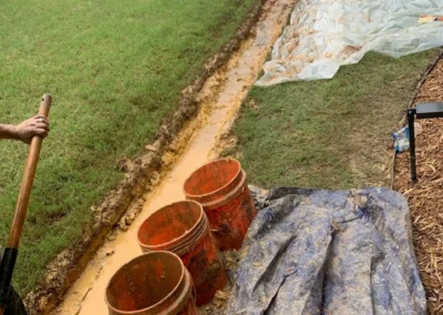 A muddy trench in a backyard with three orange buckets and a tarp, while a person digs nearby in green grass and a fenced area