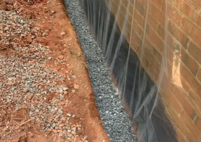 A narrow trench beside a brick wall, lined with gravel and covered by plastic sheeting to protect against moisture