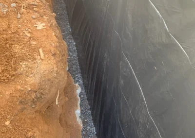 A narrow trench filled with gravel and lined with black plastic sheeting, surrounded by reddish-brown earth