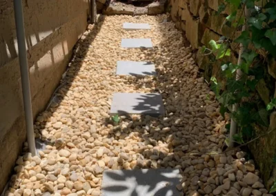 A narrow walkway lined with smooth pebbles and stepping stones, flanked by walls partially covered in greenery