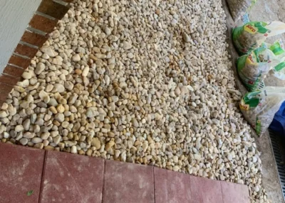A patio area featuring a path of rectangular red tiles surrounded by a layer of small, light-colored pebbles and construction materials