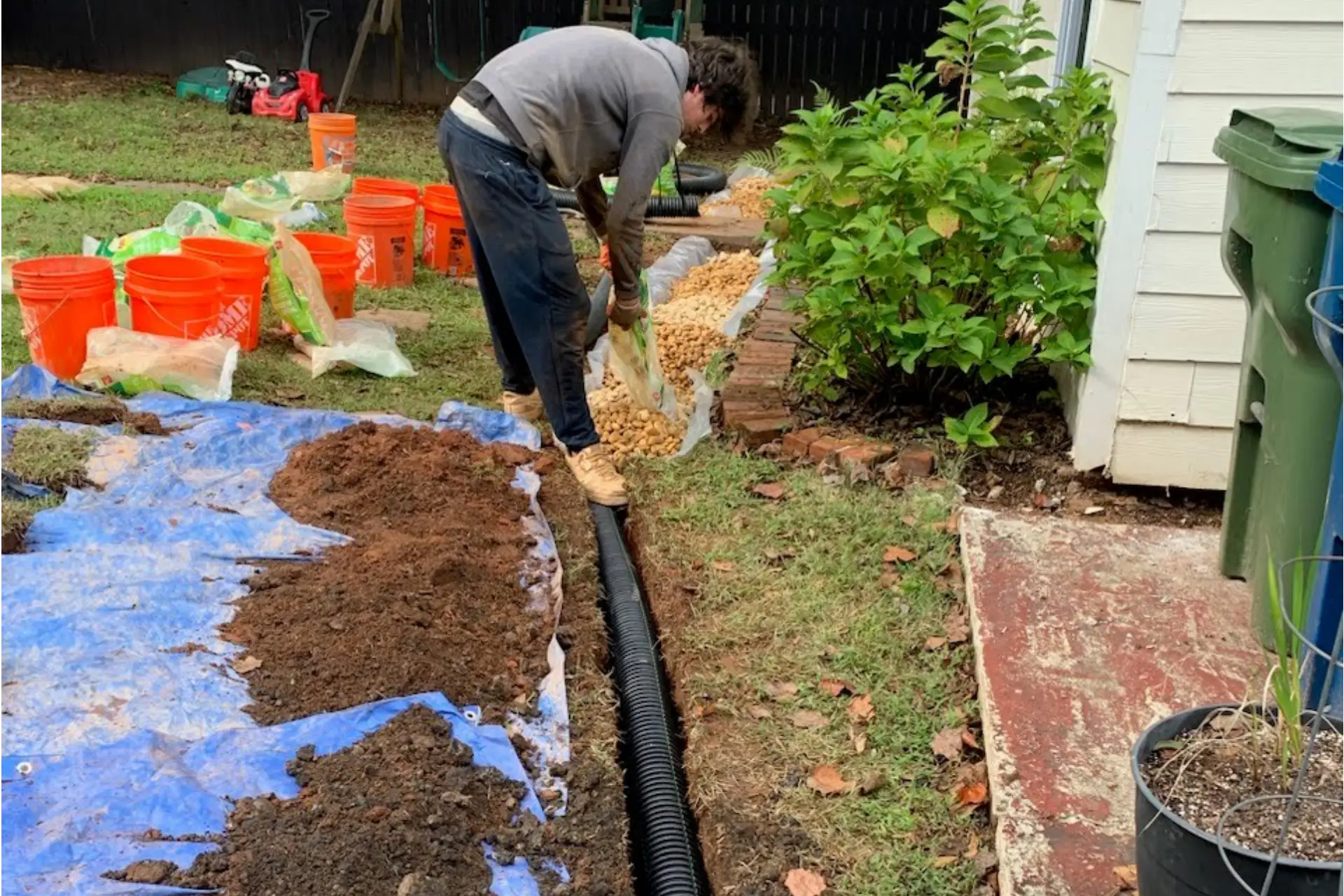 A person in a gray sweater digs along a black drainage pipe, surrounded by gardening supplies and containers in a backyard A person in a gray sweater digs along a black drainage pipe, surrounded by gardening supplies and containers in a backyard