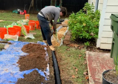 A person in a gray sweatshirt adds gravel to a trench, surrounded by orange buckets, plants, and garden tools in a backyard