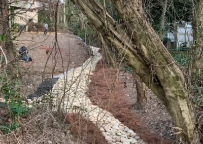 A winding pathway of smooth stones surrounded by trees and greenery, with a person working in the background
