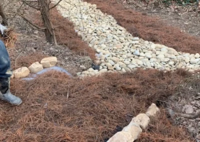 A worker constructs a stone path in a wooded area, bordered by pine needles and gravel, with greenery and a building in the background