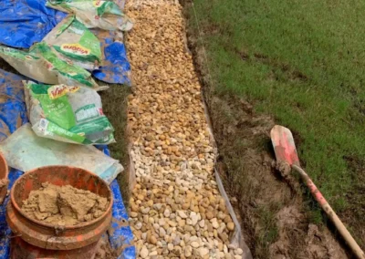 A worker places gravel along a garden path, surrounded by bags of soil and tools, with green grass in the background