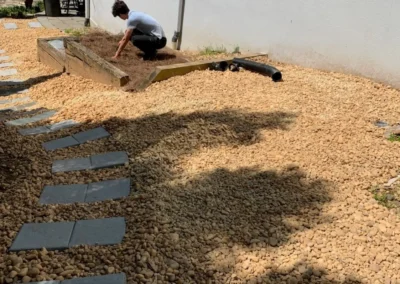 Person kneeling in a gravel-covered yard, working in a wooden planter box beside a white wall, with a stone path extending into the scene