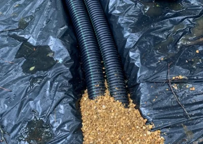 Two black drainage pipes lay on a plastic sheet, with gravel spilling out beside them in a wet, muddy setting
