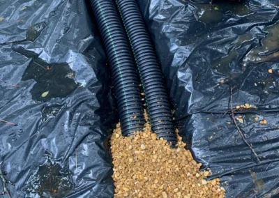 Two black drainage pipes on a black tarp, with a pile of small gravel spilling from one pipe into a puddle nearby