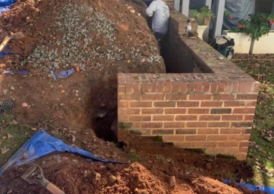 Workers excavate a large dirt pile near a brick structure, preparing for drainage or construction, surrounded by tarps and tools